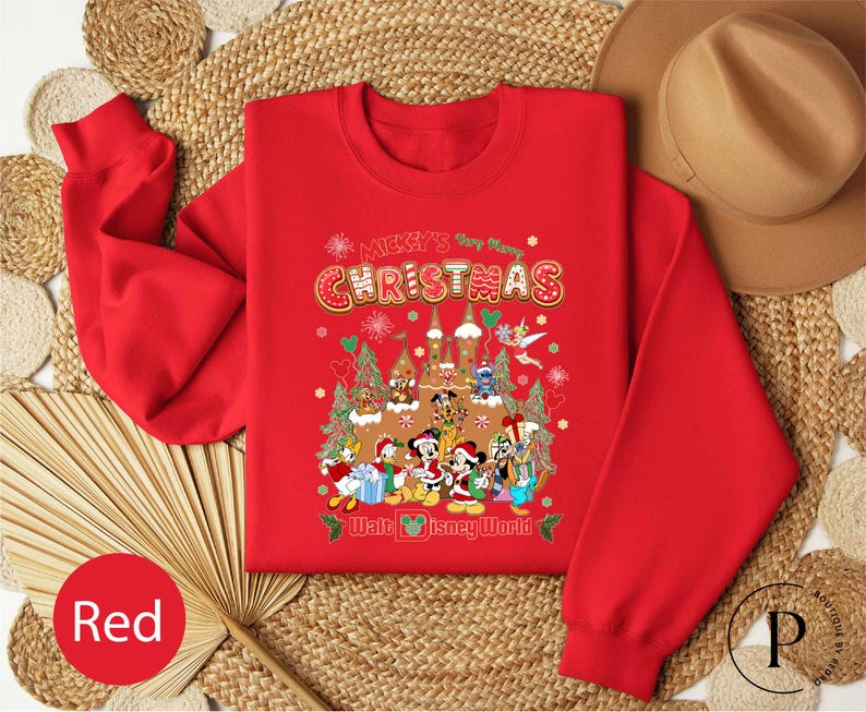 A red Christmas-themed sweatshirt with a cartoon image of Disney characters is displayed on a woven mat, along with a brown hat and a red fan.