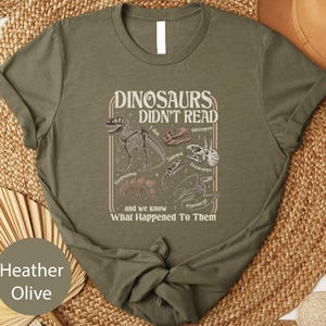 May include: Heather olive green t-shirt with a dinosaur-themed graphic. The graphic includes dinosaur skeletons and the text "DINOSAURS DIDN'T READ and we know What Happened To Them".