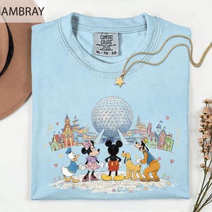 A light blue t-shirt with a Disney-themed graphic of Mickey Mouse, Minnie Mouse, Donald Duck, Goofy, and Pluto, along with a globe and various landmarks, is displayed on a white surface.