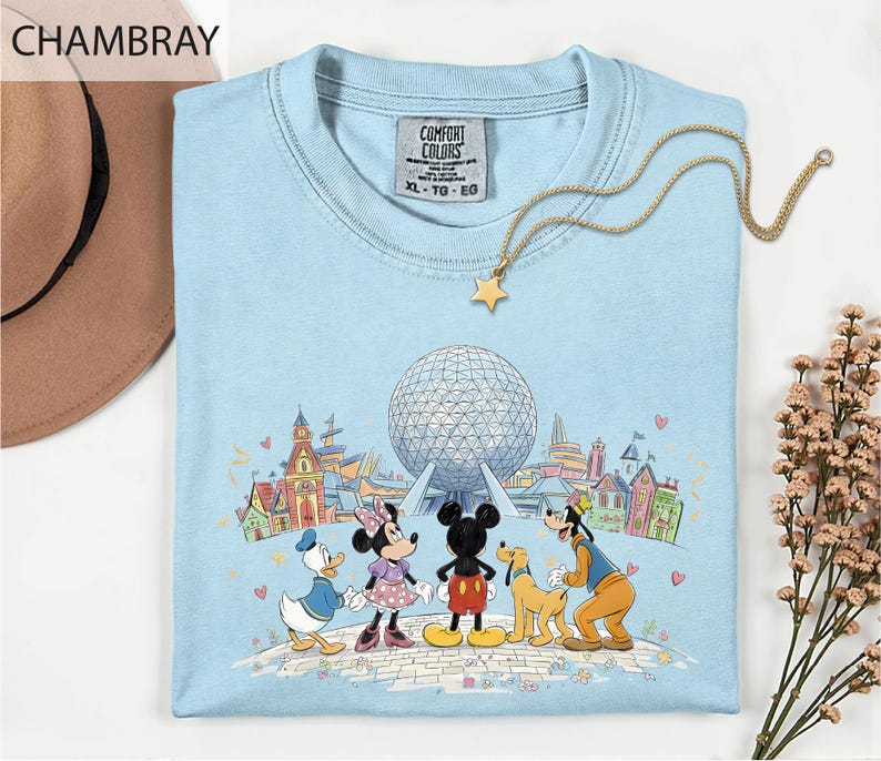 A light blue t-shirt with a Disney-themed graphic of Mickey Mouse, Minnie Mouse, Donald Duck, Goofy, and Pluto, along with a globe and various landmarks, is displayed on a white surface.