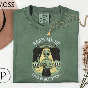 May include: Moss green Comfort Colors t-shirt with the text "BEAM ME UP THIS PLACE SUCKS" and an alien graphic. The alien is holding a drink and giving a peace sign. A gold star necklace is draped on the shirt.