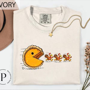 May include: Ivory-colored t-shirt with a humorous graphic of a pie chasing turkeys, inspired by the Pac-Man game. The shirt has a crew neck and short sleeves. The tag reads "Comfort Colors". A gold necklace with a star pendant is also visible.