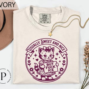 a white t-shirt with a graphic design of a cat holding a sign that says "BUT FUCK ICE" in purple text. The t-shirt is laid out on a white surface, with a hat and some flowers visible in the background.