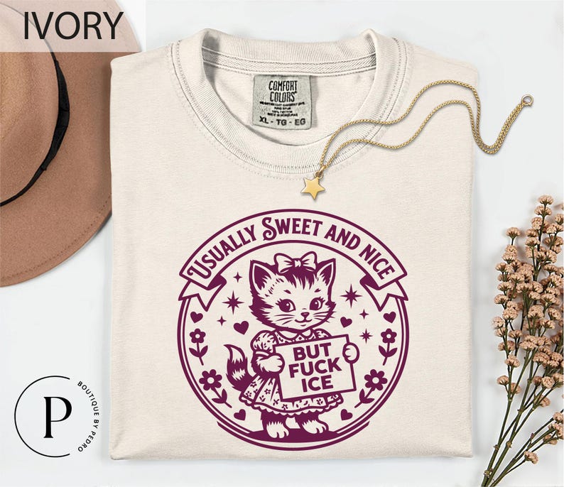a white t-shirt with a graphic design of a cat holding a sign that says "BUT FUCK ICE" in purple text. The t-shirt is laid out on a white surface, with a hat and some flowers visible in the background.