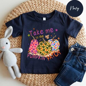 A navy blue t-shirt with a pumpkin patch design and the text "Take me to the pumpkin patch" printed on it, along with a stuffed rabbit and a pair of jeans.