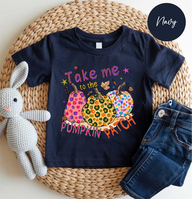 A navy blue t-shirt with a pumpkin patch design and the text "Take me to the pumpkin patch" printed on it, along with a stuffed rabbit and a pair of jeans.