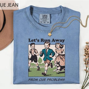 May include: A blue jean colored t-shirt featuring a graphic print of three children and a dog running in a field. The text above the graphic reads Let's Run Away and the text below reads FROM OUR PROBLEMS. The shirt has a Comfort Colors label.