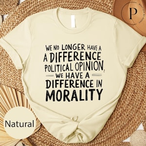 May include: A natural-colored t-shirt with the text "WE NO LONGER HAVE A DIFFERENCE POLITICAL OPINION. WE HAVE A DIFFERENCE IN MORALITY" in black. The shirt is knotted at the bottom.