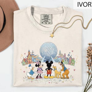 A white t-shirt with a Disney-themed graphic of Mickey Mouse, Minnie Mouse, Goofy, and Donald Duck standing in front of the Earth's Fairground, with a globe and various landmarks in the background.