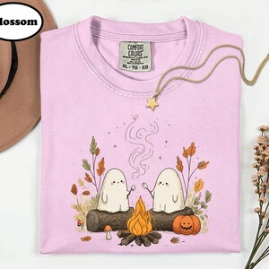 A pink t-shirt with a graphic design depicting two ghostly figures roasting marshmallows over a campfire, surrounded by autumn foliage and a star.