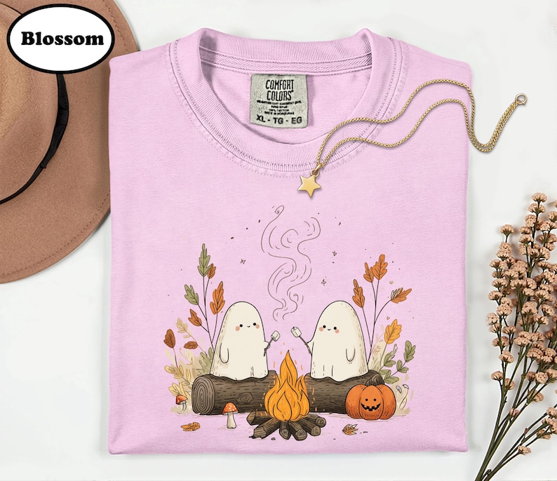 A pink t-shirt with a graphic design depicting two ghostly figures roasting marshmallows over a campfire, surrounded by autumn foliage and a star.