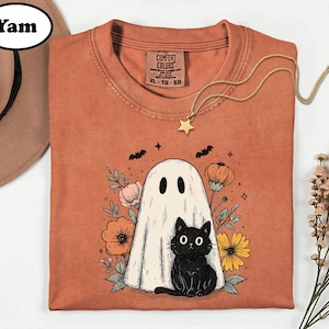 May include: A rust-colored t-shirt featuring a graphic of a white ghost and a black cat surrounded by colorful flowers, pumpkins, and bats. The shirt has a Comfort Colors label at the neck, and a gold-toned star pendant necklace rests on top.