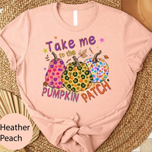 A pink t-shirt with a graphic design featuring three colorful pumpkins and the text "Take me to the PUMPKIN PATCH" printed on it.
