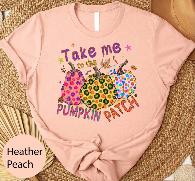 A pink t-shirt with a graphic design featuring three colorful pumpkins and the text "Take me to the PUMPKIN PATCH" printed on it.
