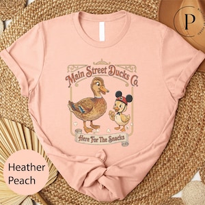 A pink t-shirt with a cartoon image of ducks and the text "Main Street Ducks Co. Here for the Snacks" printed on it.