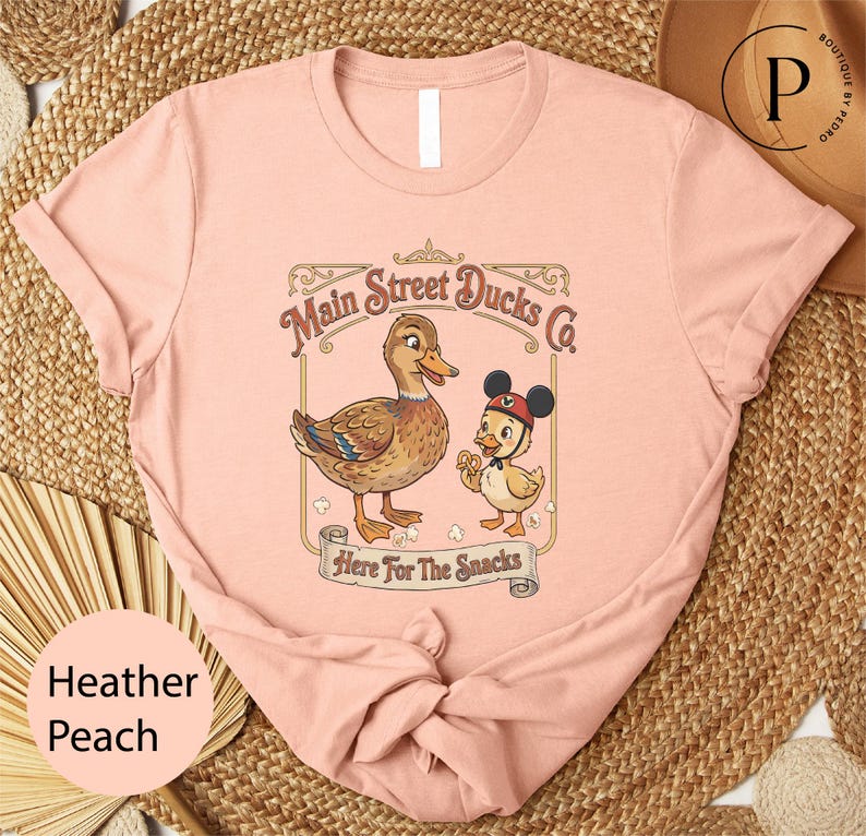 A pink t-shirt with a cartoon image of ducks and the text "Main Street Ducks Co. Here for the Snacks" printed on it.