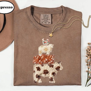 May include: An espresso-colored t-shirt featuring a stacked graphic of a chicken, sheep, pig, and cow. The animals are rendered in various fabric patterns including floral, sunburst, and cow print. The shirt has a Comfort Colors label in size XL.