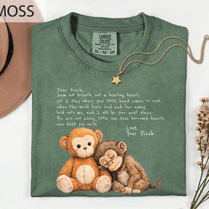 May include: Moss green Comfort Colors t-shirt with a graphic of two plush monkeys and the text "Dear Punch, I am not breath..." A gold star pendant necklace is draped on the shirt.