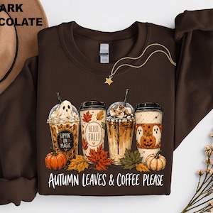 May include: A dark chocolate brown crewneck sweatshirt featuring a graphic of four coffee cups surrounded by autumn leaves and pumpkins. The cups display text reading Sippin on Magic, Hello Fall, and Autumn Leaves & Coffee Please in white lettering.