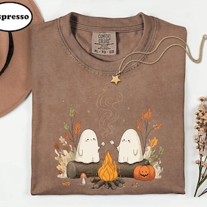 a brown t-shirt with a Halloween-themed graphic design, including two ghostly figures, a campfire, pumpkins, and autumn leaves.