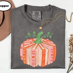 A gray t-shirt with a graphic design of a pumpkin made up of books, accompanied by a necklace and a hat.