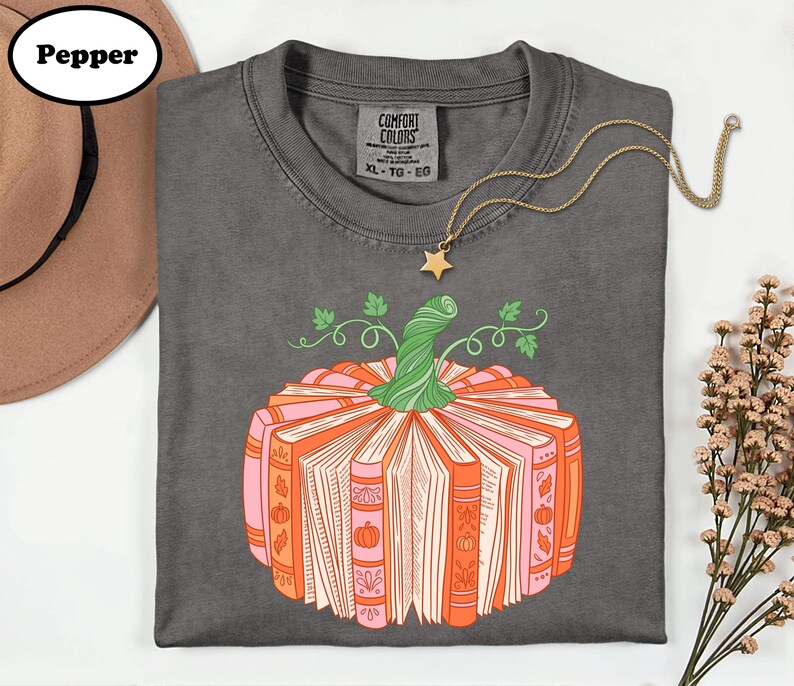 A gray t-shirt with a graphic design of a pumpkin made up of books, accompanied by a necklace and a hat.
