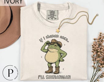 May include: Ivory-colored t-shirt with a cartoon frog wearing a cowboy hat and boots. The text reads, "If I shenan once... I'll shenanigan." A gold star pendant necklace and a brown hat are also visible.