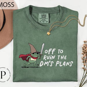 a green t-shirt with a cartoon frog wearing a wizard's hat and holding a sword, along with the text "Off to Ruin the DM's Plans" printed on it.
