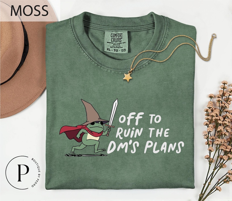 a green t-shirt with a cartoon frog wearing a wizard's hat and holding a sword, along with the text "Off to Ruin the DM's Plans" printed on it.