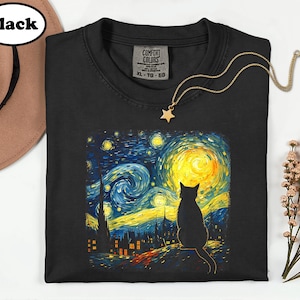 May include: A black short-sleeved t-shirt featuring a graphic print of a cat sitting before a swirling night sky with a large yellow moon. The shirt has a Comfort Colors label at the neck with size XL, TG, EG printed on it and a gold star necklace.