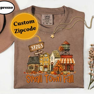 May include: An espresso-colored t-shirt featuring a graphic of a small town street scene with houses, a water tower, pumpkins, and autumn trees. The text reads 37203, Nashville, TN, and Small Town Fall. The shirt has a Comfort Colors brand label.