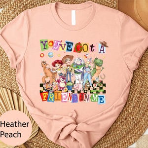 A pink t-shirt with a colorful graphic design featuring characters from the Toy Story movie, including Woody, Buzz Lightyear, and other beloved characters.