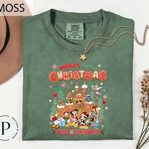 A green t-shirt with a Christmas-themed graphic design, including various Disney characters and holiday decorations, is displayed on a white background.