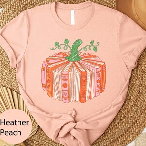 A pink t-shirt with a graphic design of a pumpkin made up of books, surrounded by leaves and vines.