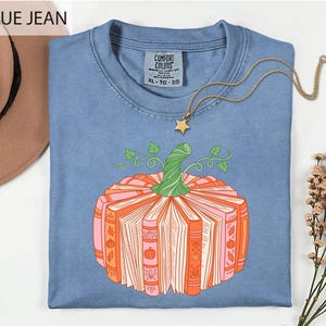 A blue t-shirt with a graphic of a pumpkin made of books, accompanied by a necklace and a hat, all placed on a white background.