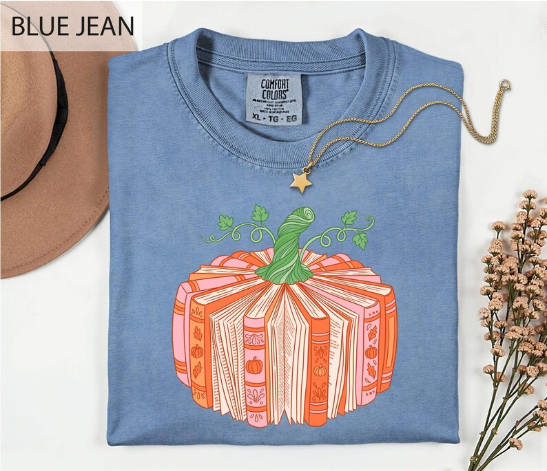A blue t-shirt with a graphic of a pumpkin made of books, accompanied by a necklace and a hat, all placed on a white background.