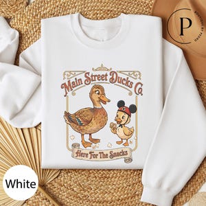 A white t-shirt with a graphic design featuring two cartoon ducks, one larger and one smaller, along with the text "Main Street Ducks Co." and "Here for the Snacks".