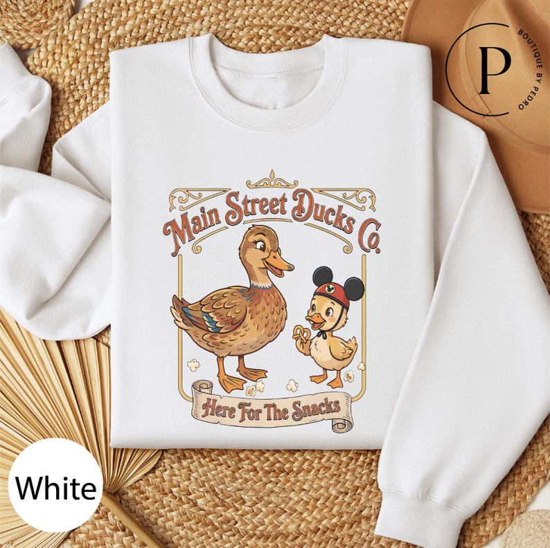 A white t-shirt with a graphic design featuring two cartoon ducks, one larger and one smaller, along with the text "Main Street Ducks Co." and "Here for the Snacks".