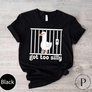 Peut inclure: T-shirt noir avec un dessin d'oie en cage et le texte "got too silly" en blanc. L'oie est blanche avec un bec et des pattes oranges. Le t-shirt a un col rond et des manches courtes.