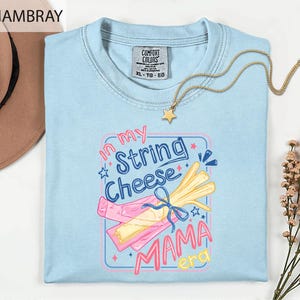 May include: Light blue t-shirt with the text "in my String Cheese Mama era" in a colorful graphic. The shirt features a crew neck and a tag that says "Comfort Colors". A gold necklace with a star pendant is draped on the shirt.