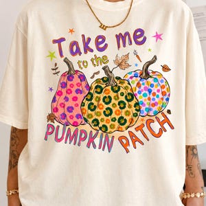 A person wearing a white t-shirt with a colorful graphic design featuring three pumpkins and the text "Take me to the pumpkin patch".
