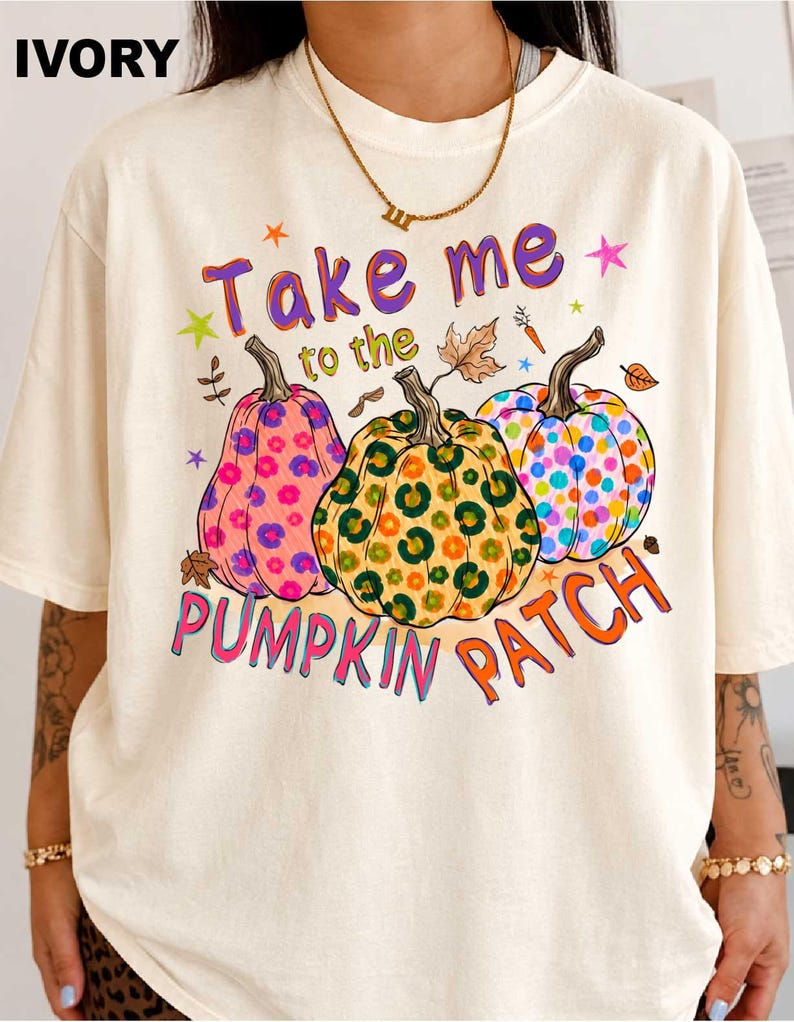 A person wearing a white t-shirt with a colorful graphic design featuring three pumpkins and the text "Take me to the pumpkin patch".