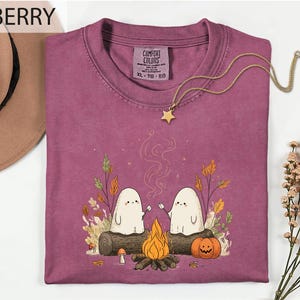 a purple t-shirt with a graphic design depicting two ghostly figures sitting by a campfire, surrounded by autumn foliage and a pumpkin. The t-shirt is displayed on a white background, with a hat and some flowers visible in the background.