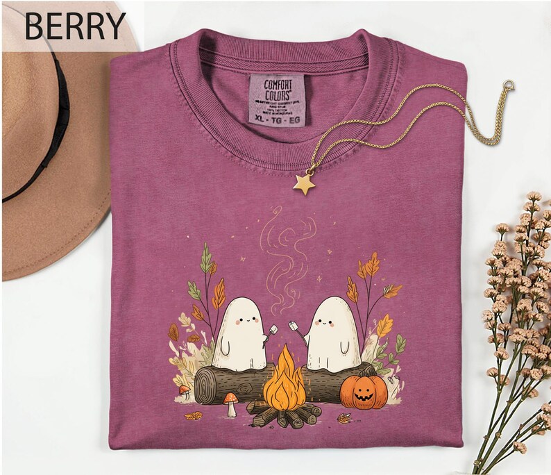 a purple t-shirt with a graphic design depicting two ghostly figures sitting by a campfire, surrounded by autumn foliage and a pumpkin. The t-shirt is displayed on a white background, with a hat and some flowers visible in the background.