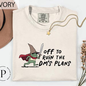 May include: Ivory-colored t-shirt with a cartoon frog wearing a wizard hat and cape, holding a sword. The text "off to ruin the DM's plans" is printed on the shirt. A gold necklace with a star pendant is also visible.