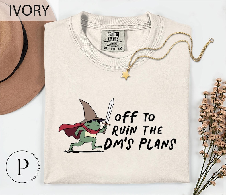 a white t-shirt with a cartoon frog wearing a wizard's hat and holding a sword, along with the text "Off to Ruin the DM's Plans".