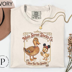 a white t-shirt with a graphic design of two cartoon ducks, one larger and one smaller, along with the text "Main Street Ducks Co." and "Here For The Snacks" printed on it. The t-shirt is displayed on a white background, with a hat and some flowers visible in the background.