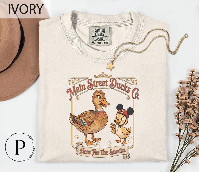 a white t-shirt with a graphic design of two cartoon ducks, one larger and one smaller, along with the text "Main Street Ducks Co." and "Here For The Snacks" printed on it. The t-shirt is displayed on a white background, with a hat and some flowers visible in the background.