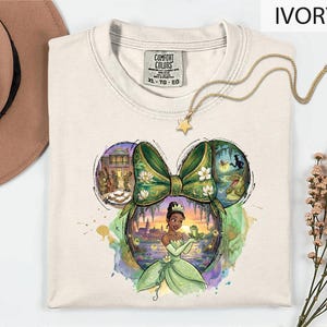 A white t-shirt with a colorful illustration of a character from the Disney film " princess and the frog" displayed on it, along with a star-shaped charm.