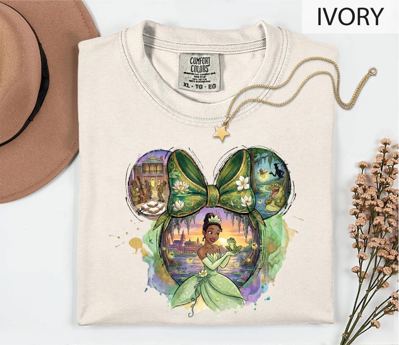 A white t-shirt with a colorful illustration of a character from the Disney film " princess and the frog" displayed on it, along with a star-shaped charm.